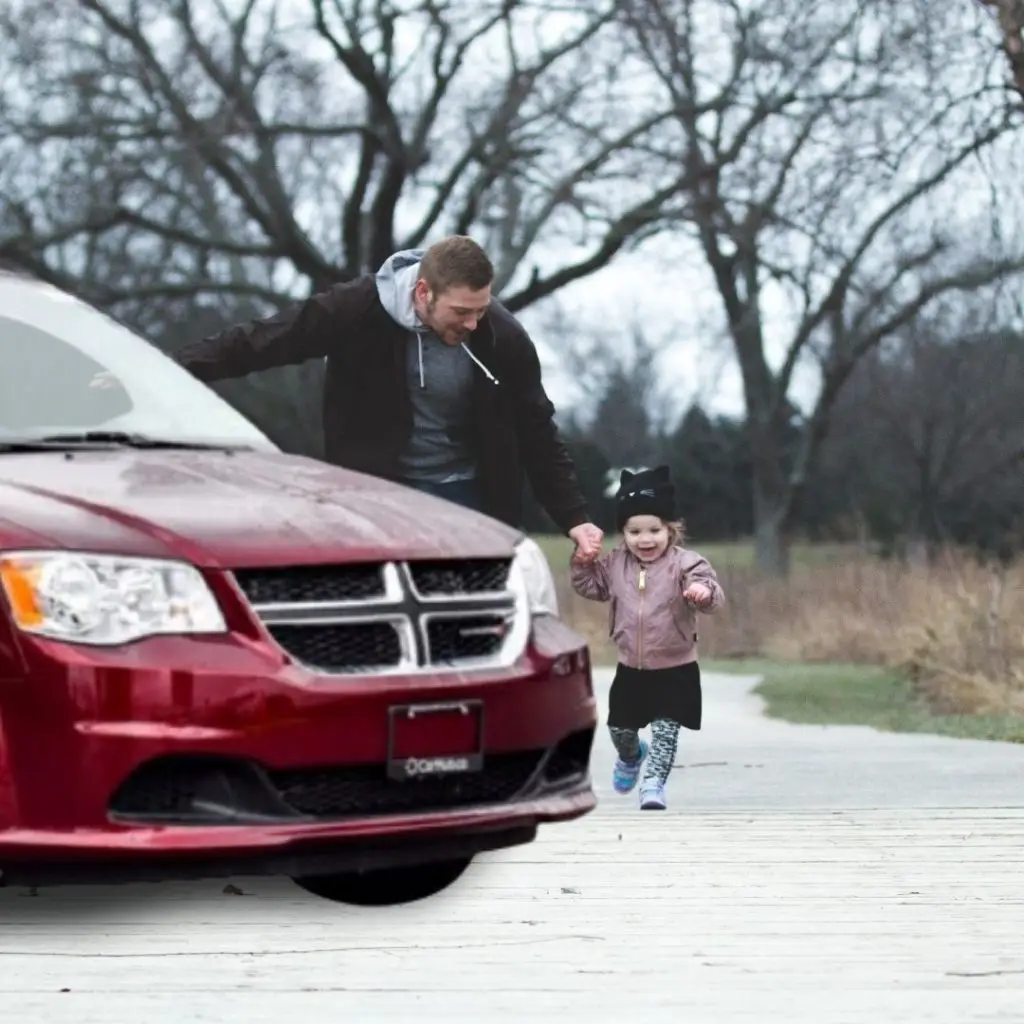 Man and toddler girl holding hands walking toward a red Dodge Grand Caravan minivan