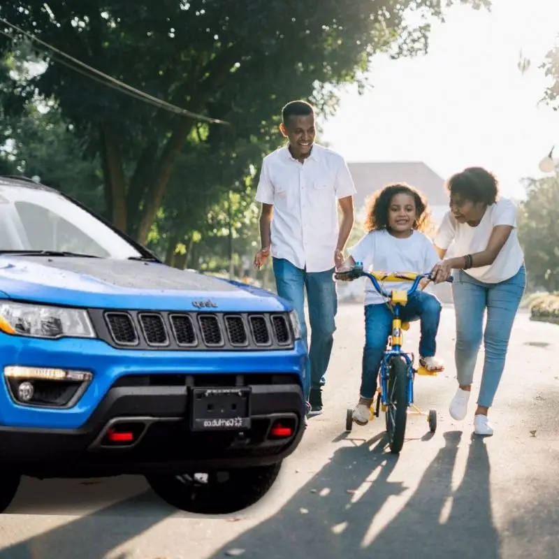 Blue Jeep Compass Trailhawk SUV with family teaching child to ride bicycle nearby