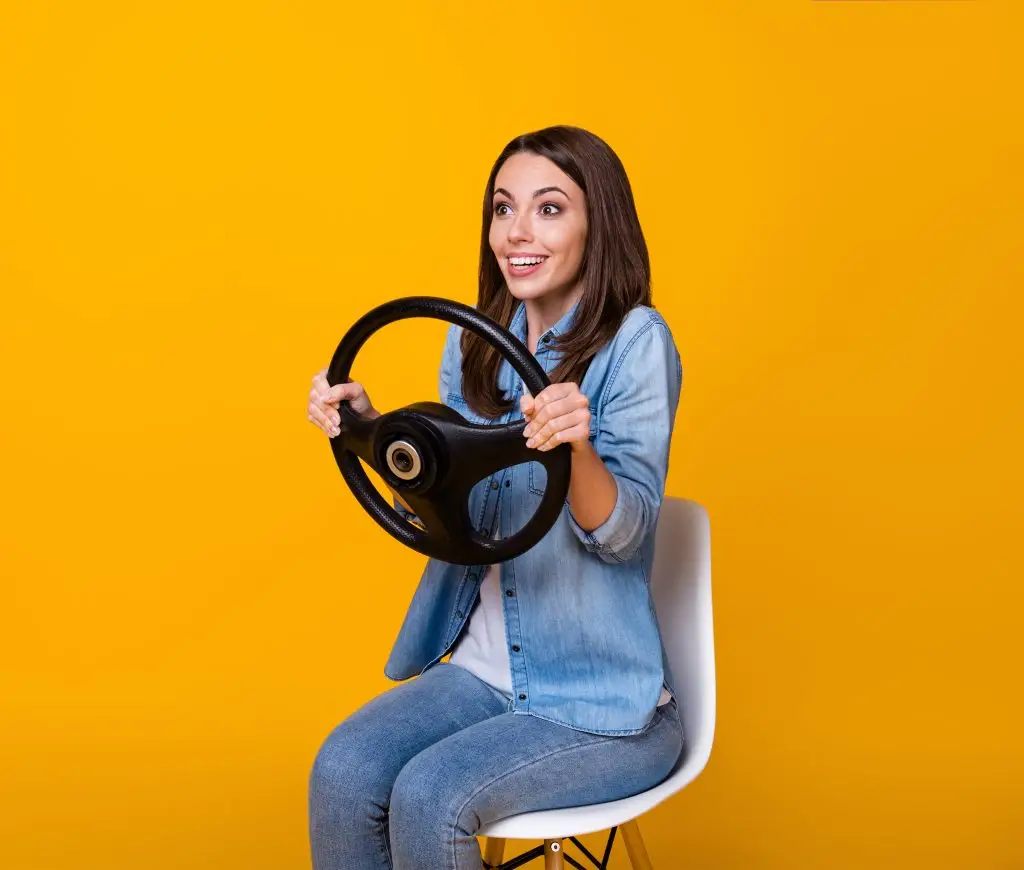 Woman sitting on chair holding car steering wheel against yellow background, playful driving concept