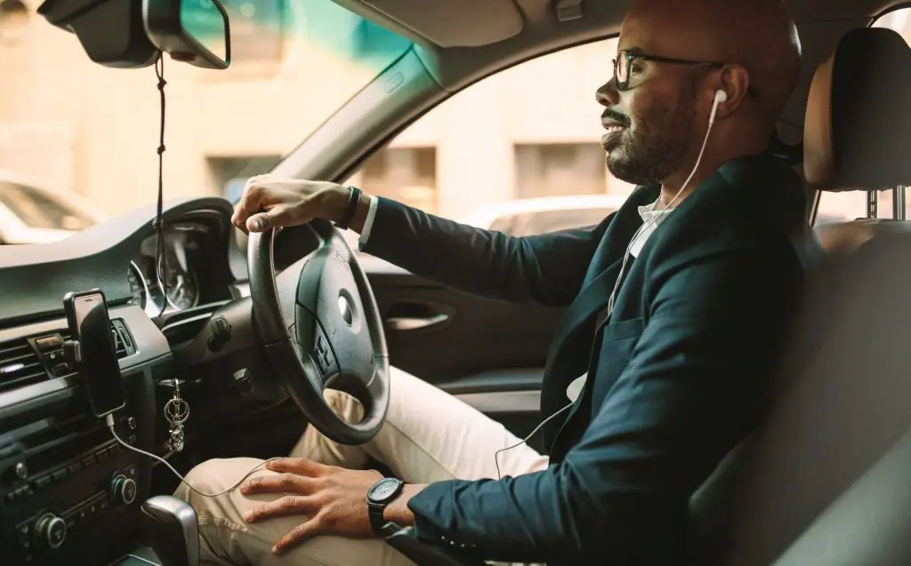 Man in business suit driving car with wireless earbuds and phone mounted on dashboard