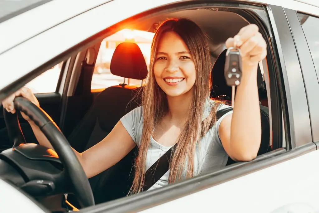 Smiling young woman holding car keys out driver's side window after buying new car