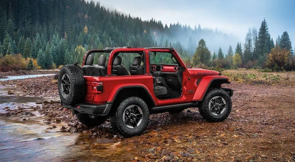 Red two-door Jeep Wrangler Rubicon parked beside forest stream with pine trees