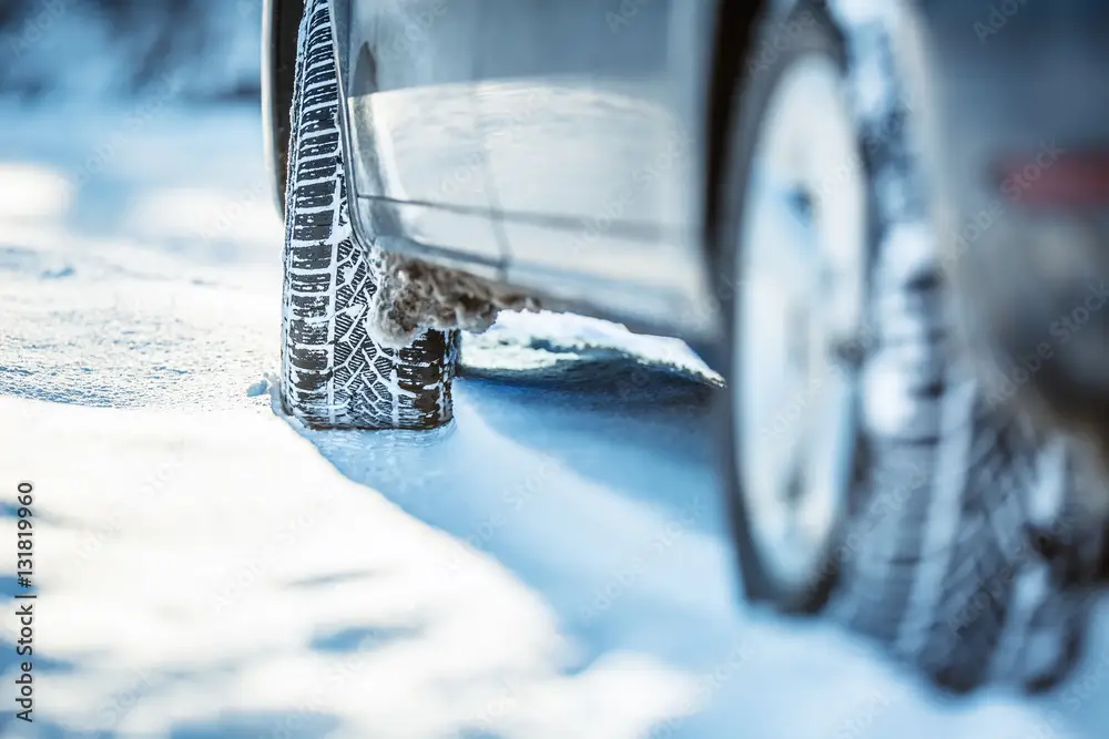 Close-up of a snow-covered tire against a vehicle parked on an icy winter road