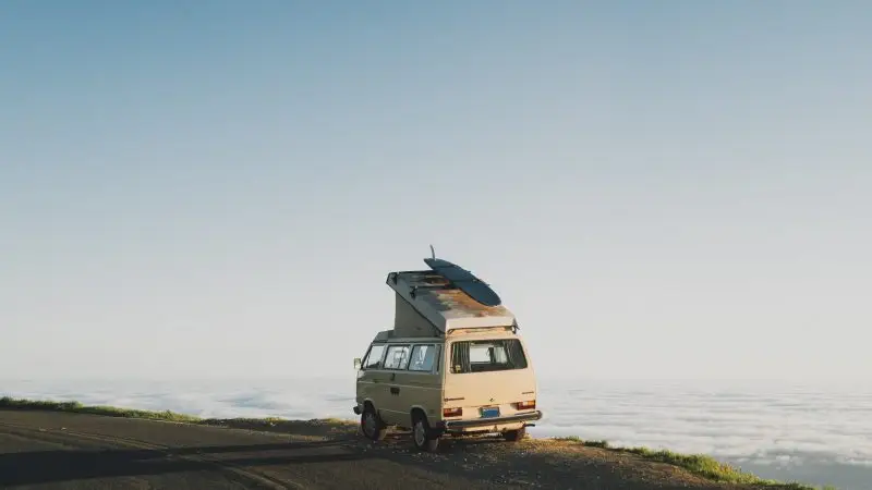 Vintage camper van with a surfboard on the roof parked on a cliff overlooking a sea of clouds