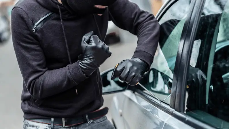 Masked thief in black gloves using a tool to break into a car through the window