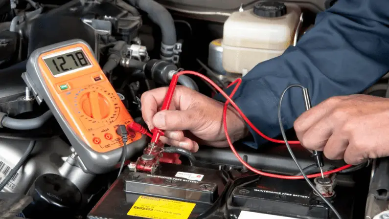 A mechanic using an orange multimeter showing 12.78 volts while testing a car battery terminal under the hood.
