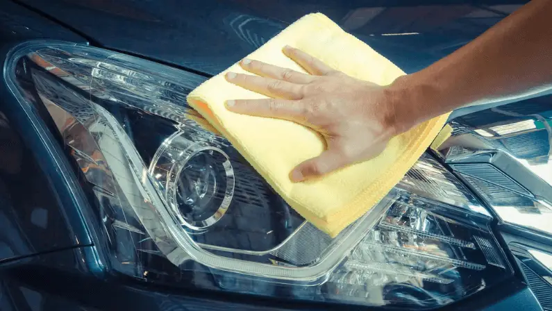A hand wiping a car's clear headlight lens with a yellow microfiber cloth.
