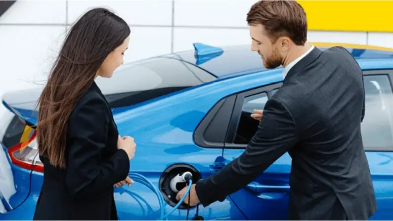 Man in a business suit plugging a charging cable into a blue electric car while a woman looks on, both standing beside charging station equipment.