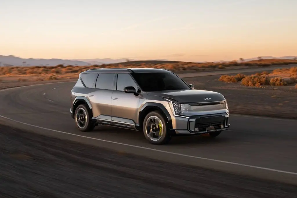Silver Kia EV9 electric SUV driving on desert highway road at sunset